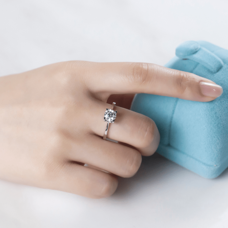 Hand with The Estelle Moissanite solitaire engagement ring resting on a blue ring box 💍✨ - an amazing value for weddings!