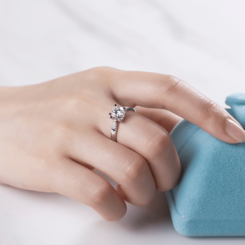Hand wearing The Seraphine Moissanite engagement ring, showcasing its brilliance next to a soft blue box 💍✨
